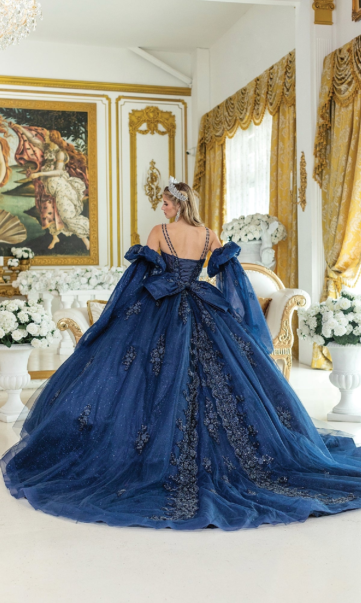 A woman in the Blue Quinceanera Dress 5037 By Dancing Queen and a tiara stands in an ornate, gold-accented room with white flowers, facing away from the camera.